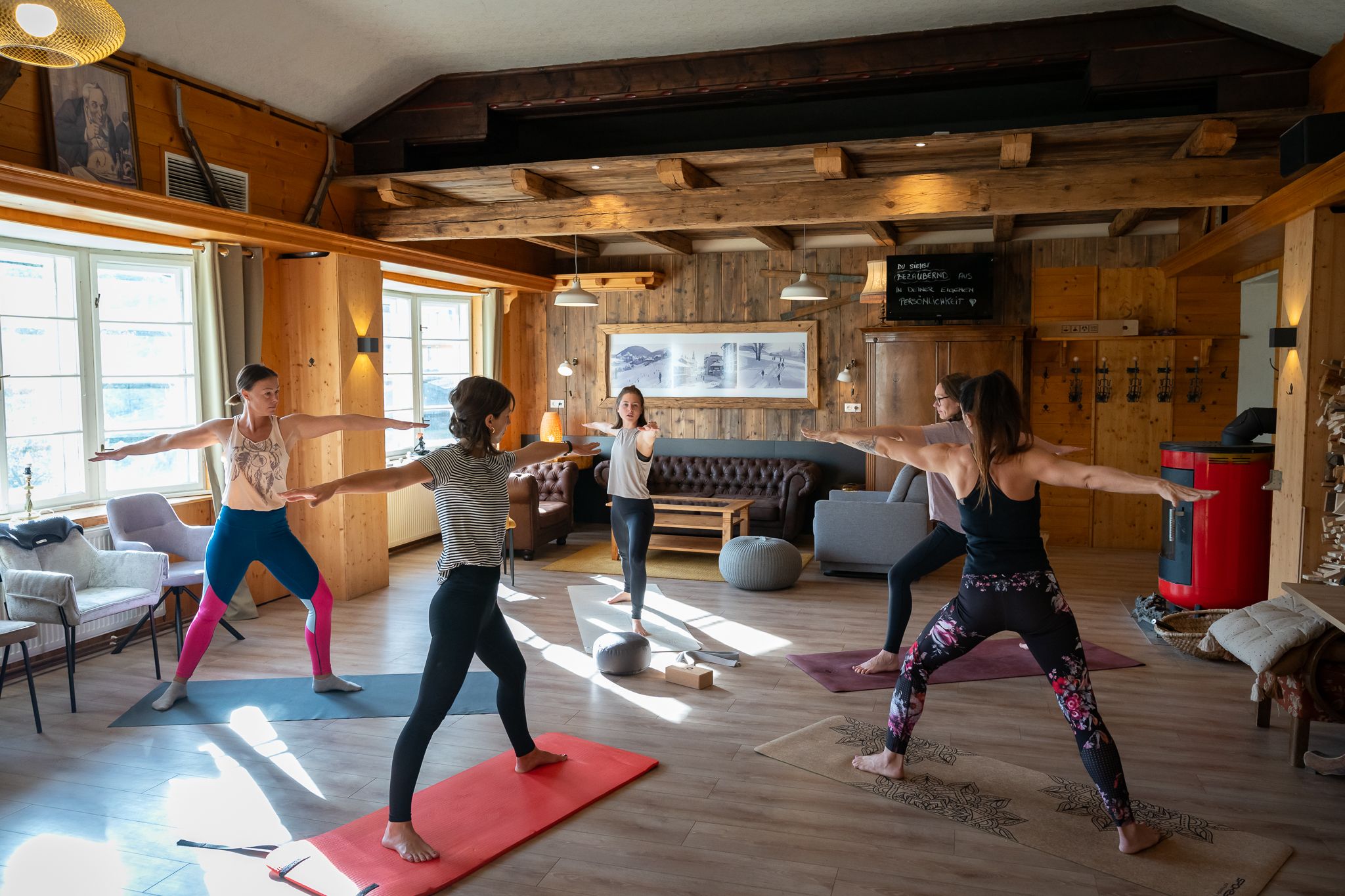 A group of women is practicing yoga in a cozy room with wooden walls and large windows.