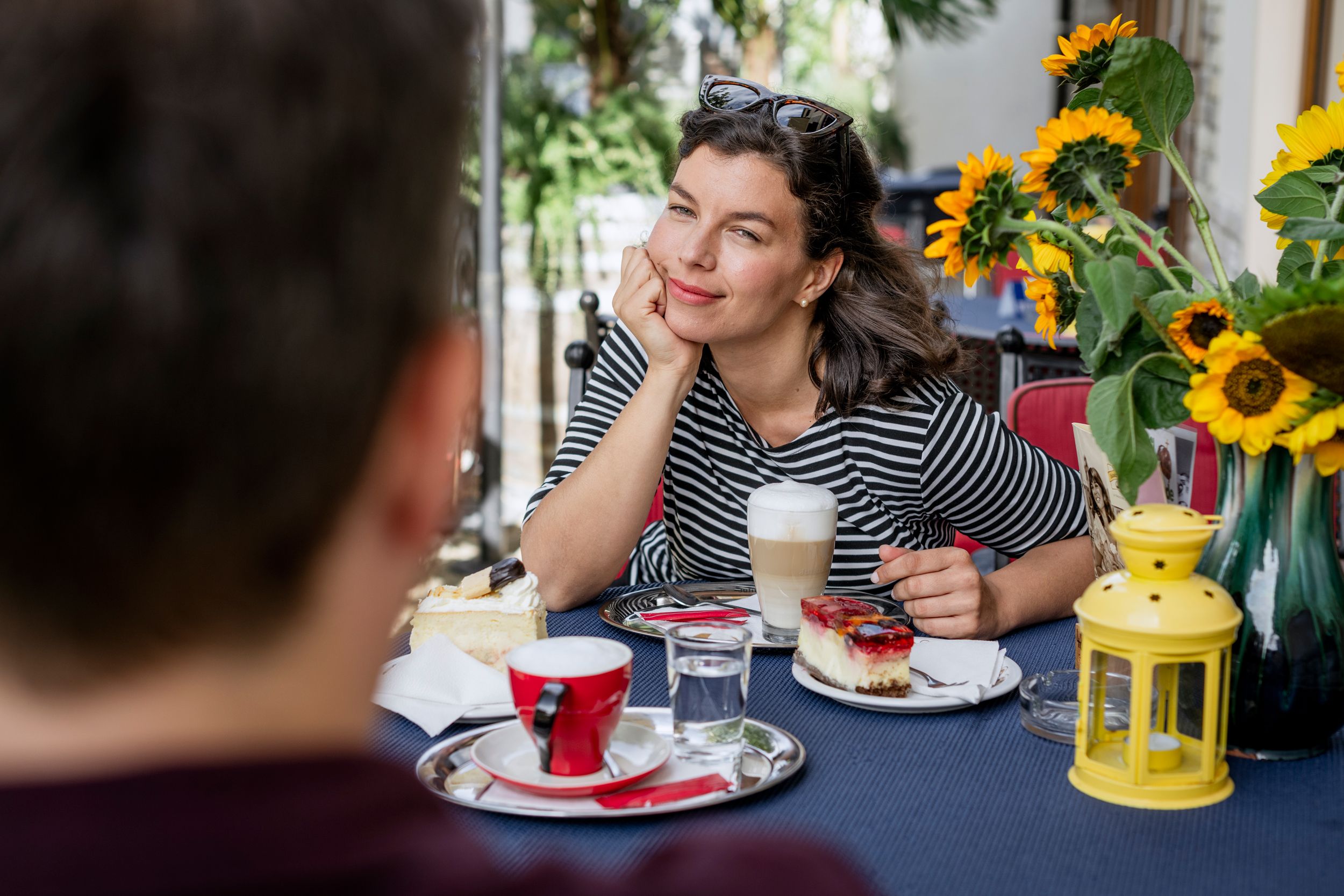 Woman in striped shirt enjoying coffee and cake outdoors, surrounded by sunflowers.