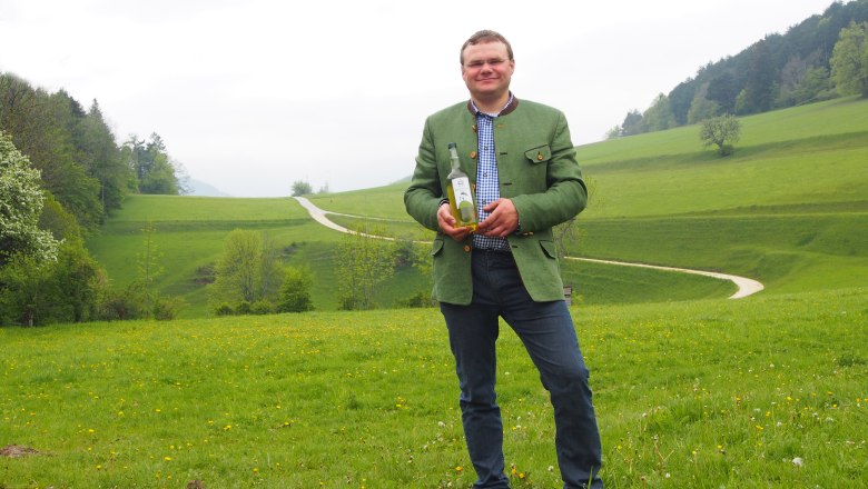 Man in green jacket holding a bottle on a green meadow with hills in the background.