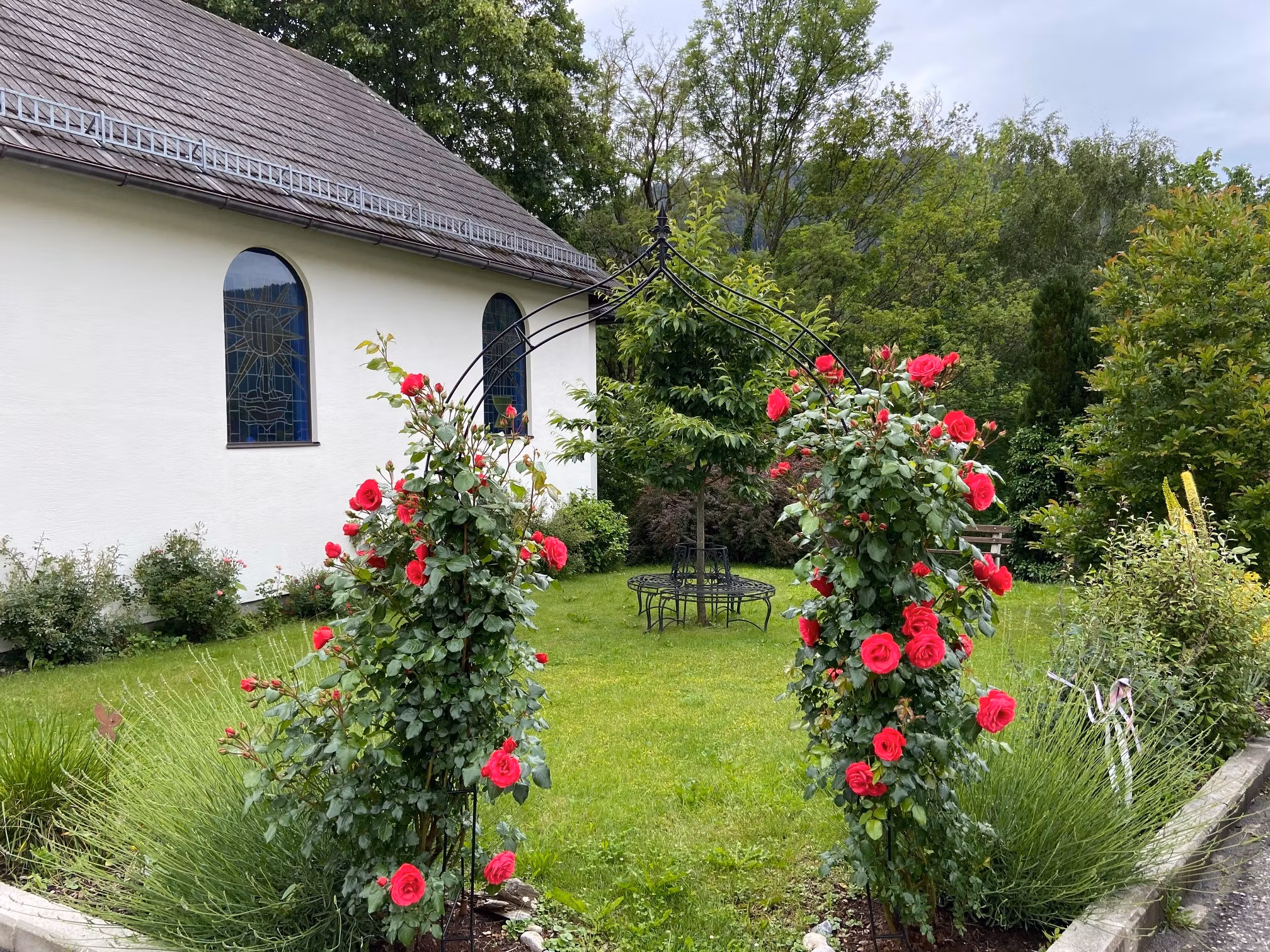 A rose arch with red roses in front of a chapel with colorful windows and a green garden.