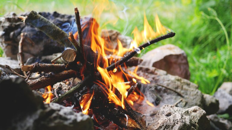 Close-up of a campfire with burning logs and flames, surrounded by stones in the open air.