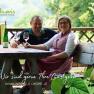 A couple sits at a wooden table outdoors with wine and glasses. Nature can be seen in the background. Text: "We are happy to be your hosts".