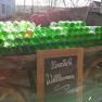 A pile of green glass bottles on a trolley with a 'Welcome' sign in front of it, a house and trees in the background.