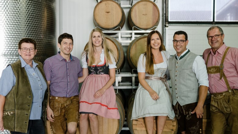 A group of six people in traditional Bavarian dress stand in front of wine barrels in a wine cellar.