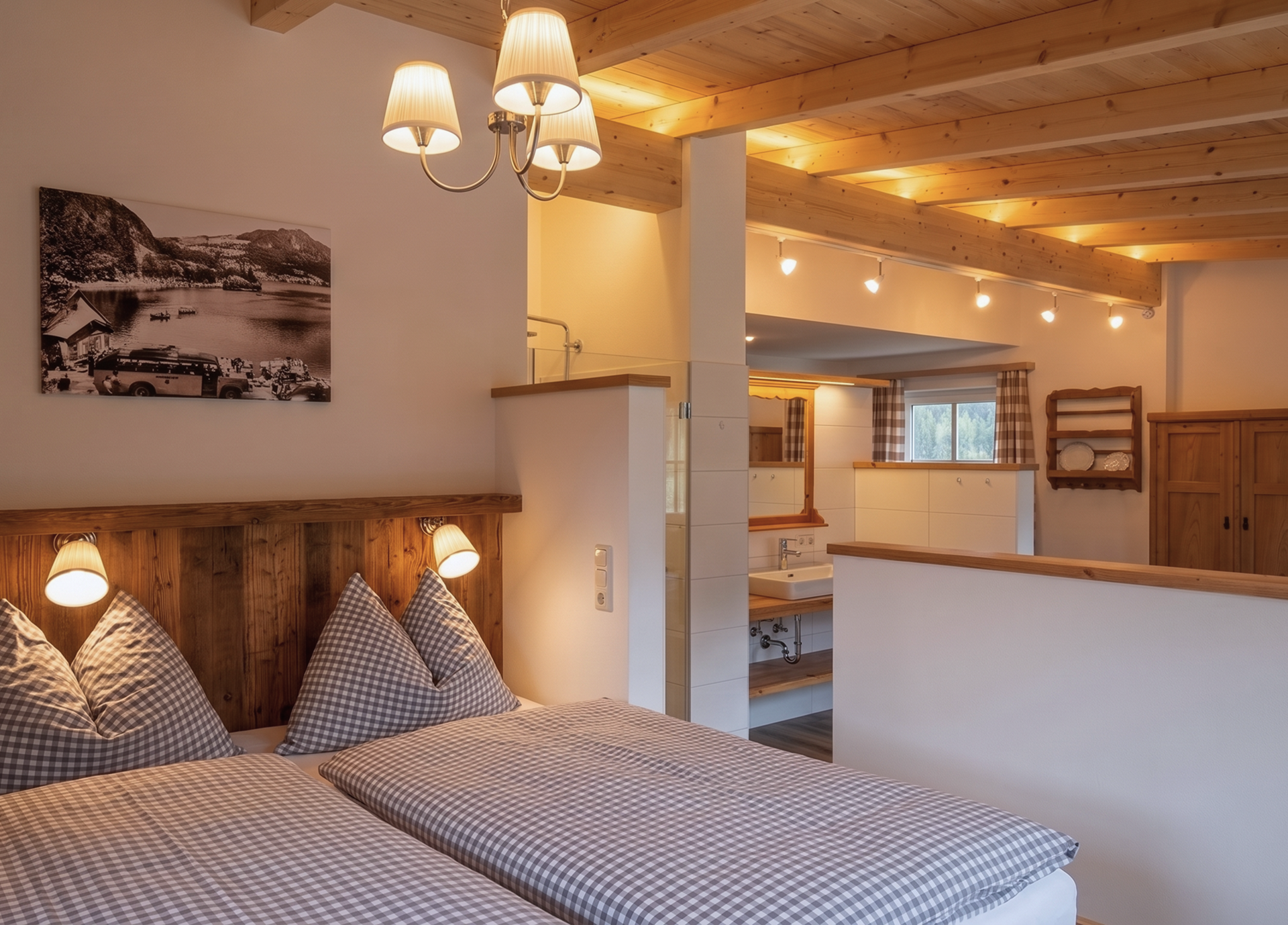 A cozy bedroom with a wooden beam ceiling and a double bed, decorated with checkered pillows and blankets. In the background is a bathroom with a sink; behind the half-height wall is a freestanding bathtub.