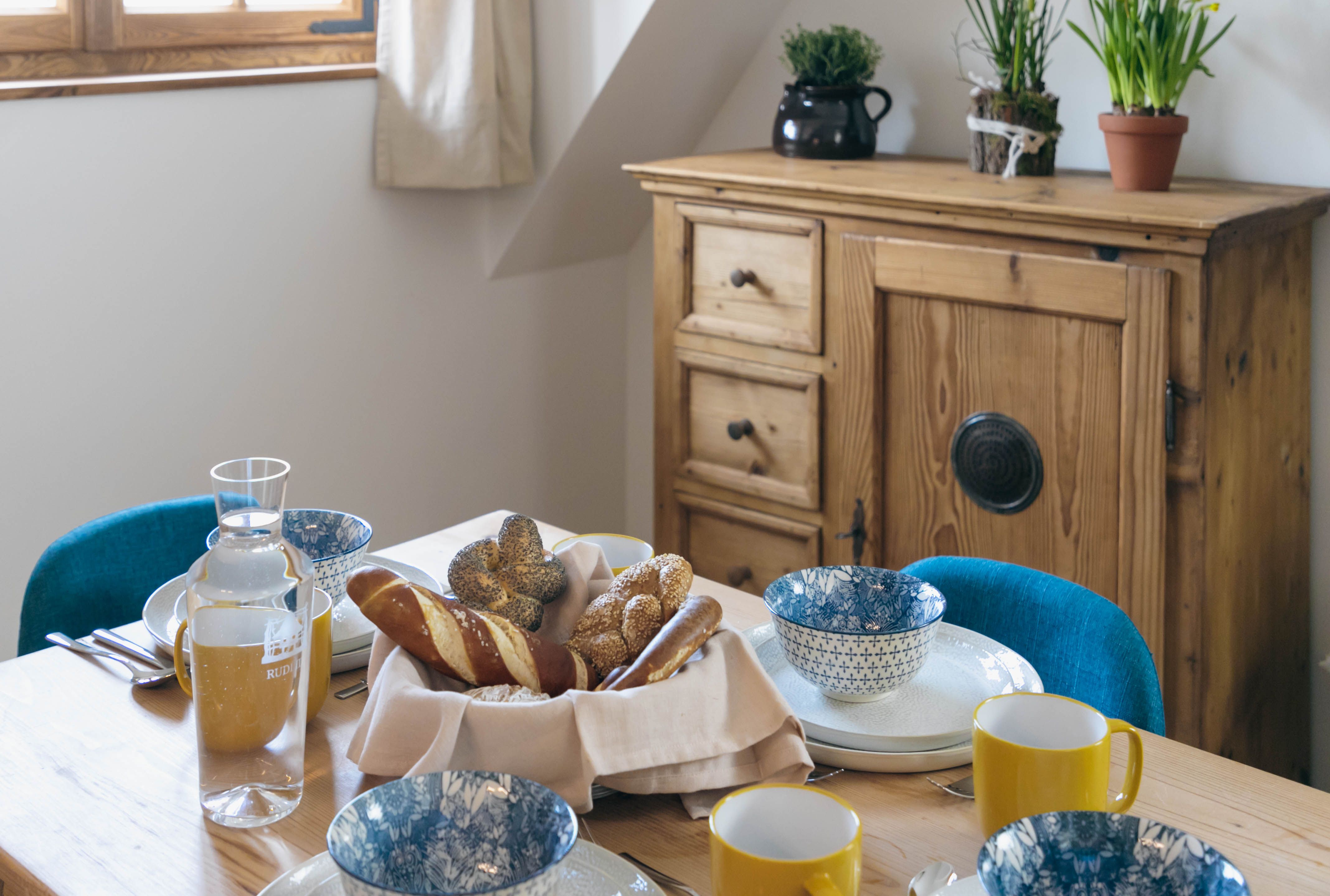 A laid breakfast table with bread rolls, crockery and yellow cups, a wooden cupboard with plants in the background.