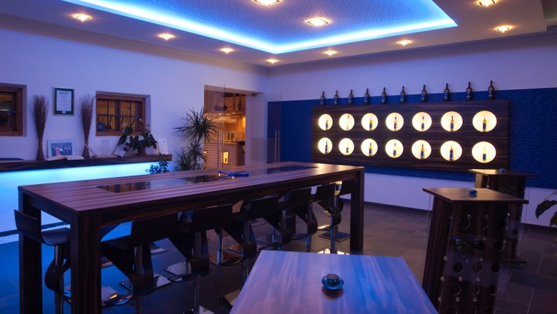 Modern tasting room with blue lighting, wooden tables and wine bottles on the wall.