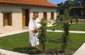 A man in antique clothing stands in a garden in front of a historic building.