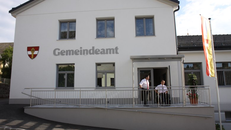 A modern municipal office with a white fa&ccedil;ade, two people standing on a ramp, a flag flying next to it.