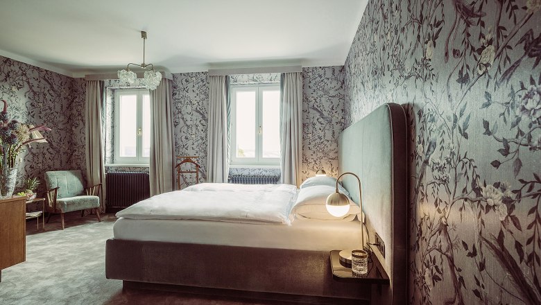 Elegant bedroom with floral patterned wallpaper, large bed and two windows.