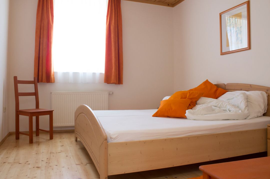 A simply furnished double room with a wooden bed, orange cushions and curtains, a chair and a picture on the wall.