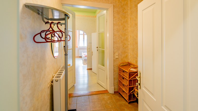 An anteroom with coat hooks, a radiator and wicker furniture. A door leads into a bright room with windows.
