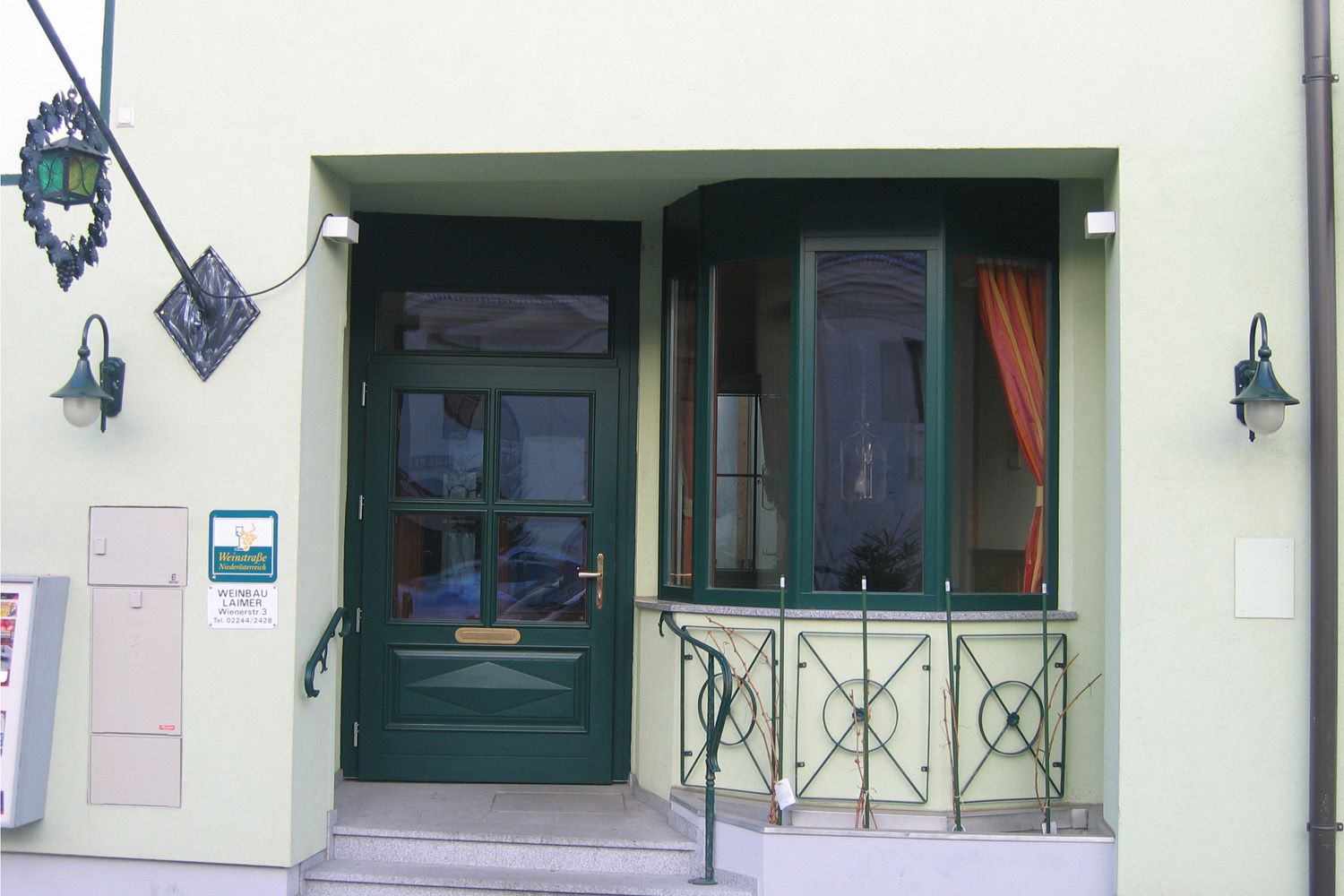 Entrance area of a building with green door and windows, decorated with plants and lanterns.