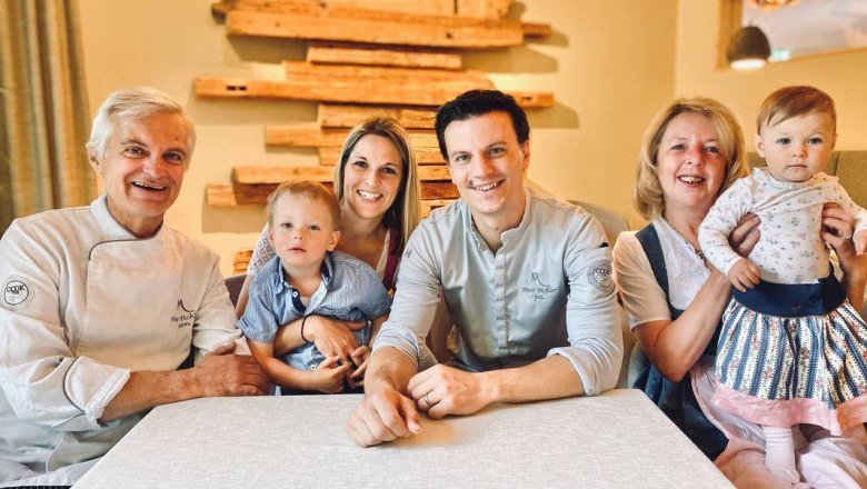 A family sits at a table and smiles at the camera. Two adults are each holding a child on their lap.