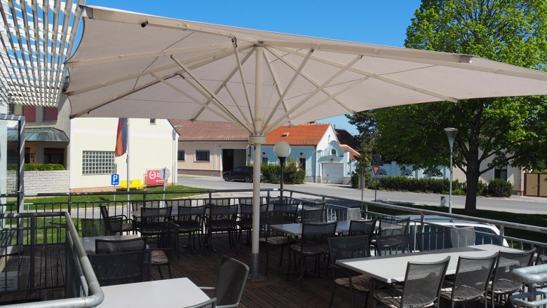 Terrace of a restaurant with parasol and empty tables and chairs, in the background a street and buildings.
