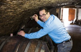 Man in blue shirt draws wine from a barrel in a wine cellar.