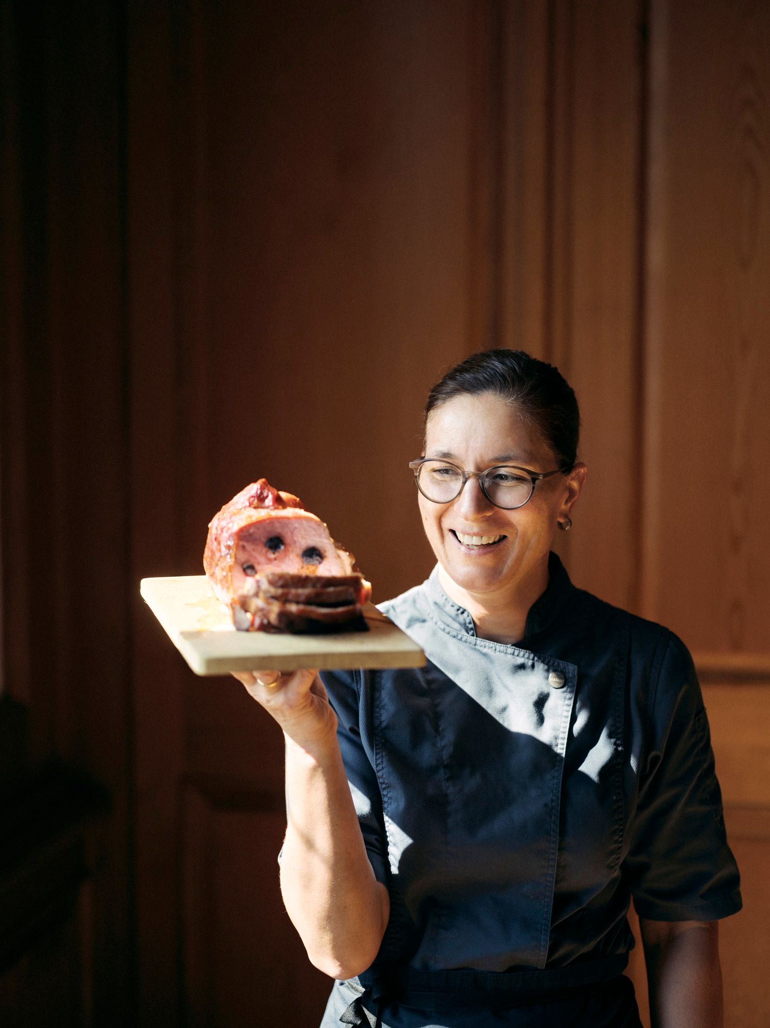 A woman in chef's clothes holds a wooden board with a sliced meat dish.