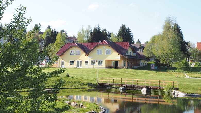 Yellow building with a red roof by a pond, surrounded by trees and lawn.