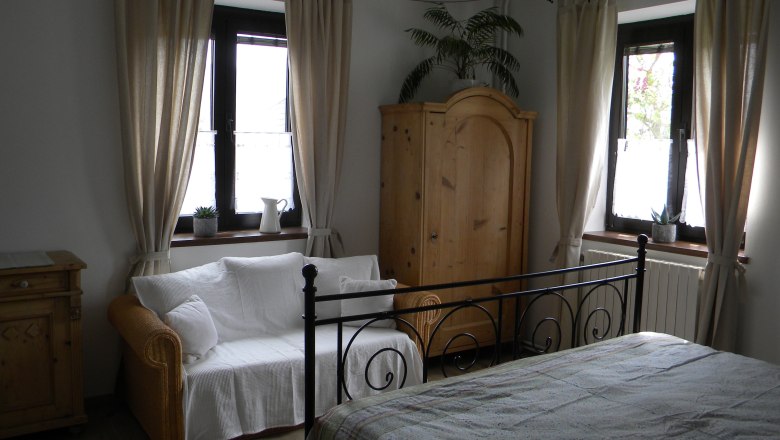 A cozy bedroom with a metal bed, a sofa, a wooden wardrobe and two windows with curtains.