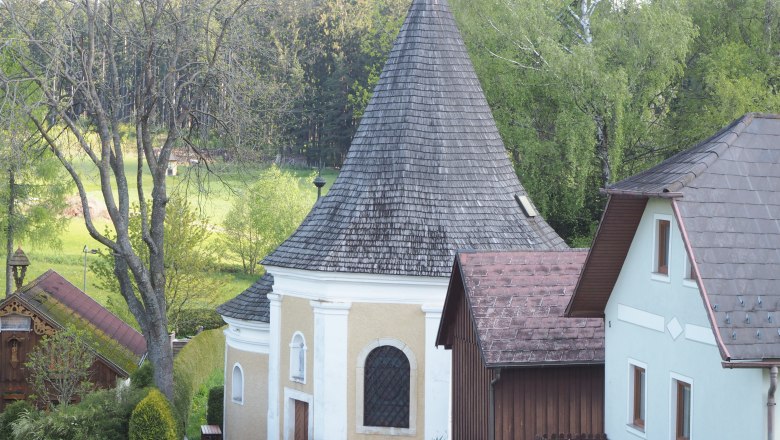 A small chapel with a pointed roof, surrounded by trees and houses.