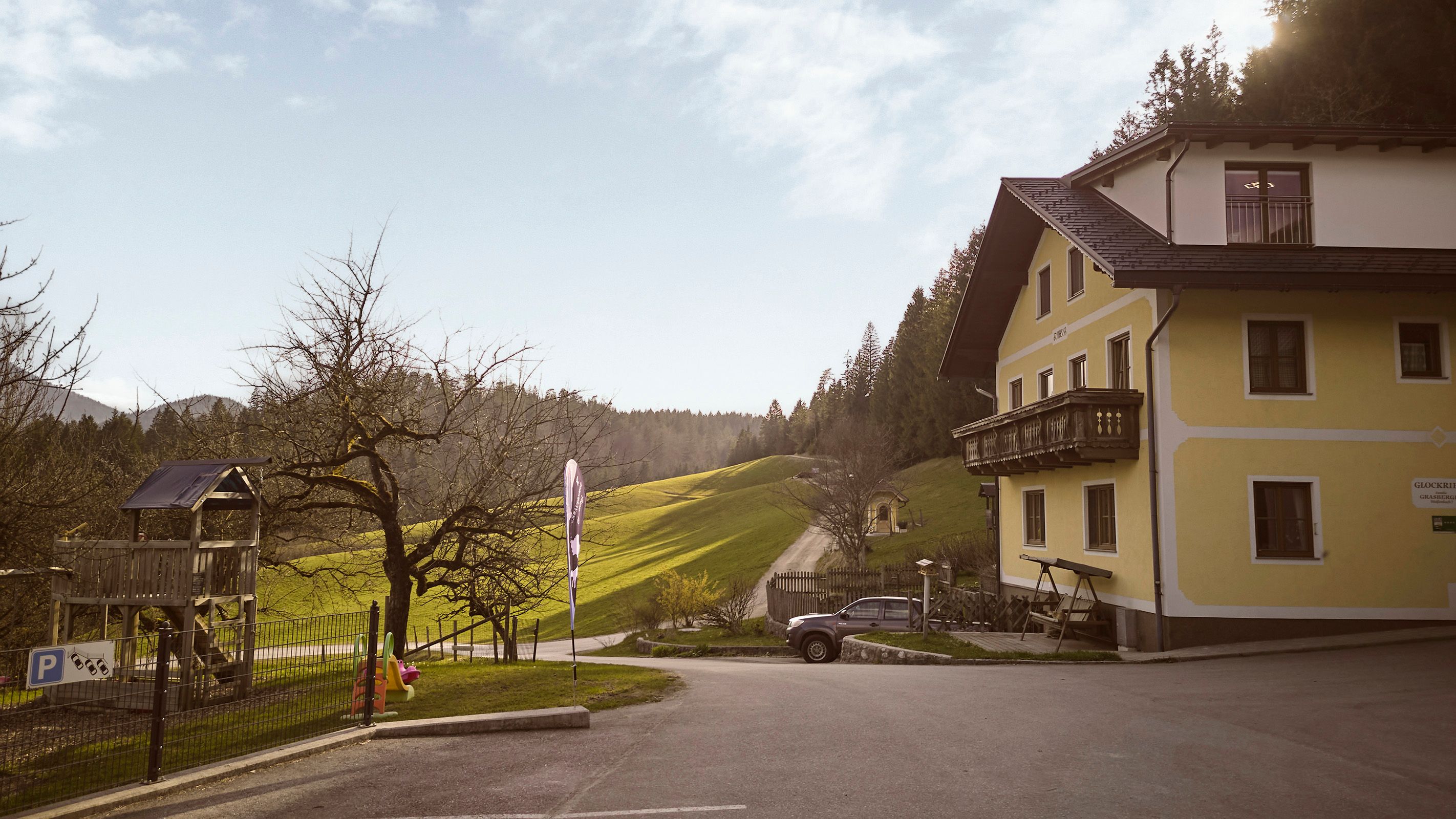 A yellow house with a balcony stands next to a playground and a road that leads through green hills. The sky is clear and the sun is shining.