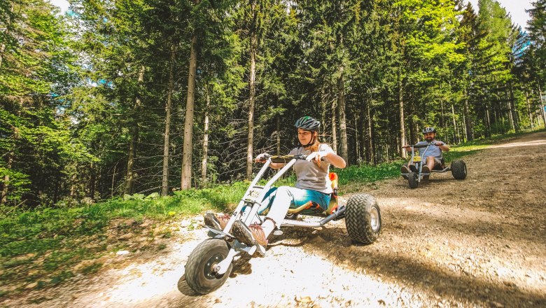 Two people are riding mountain carts on a forest path.