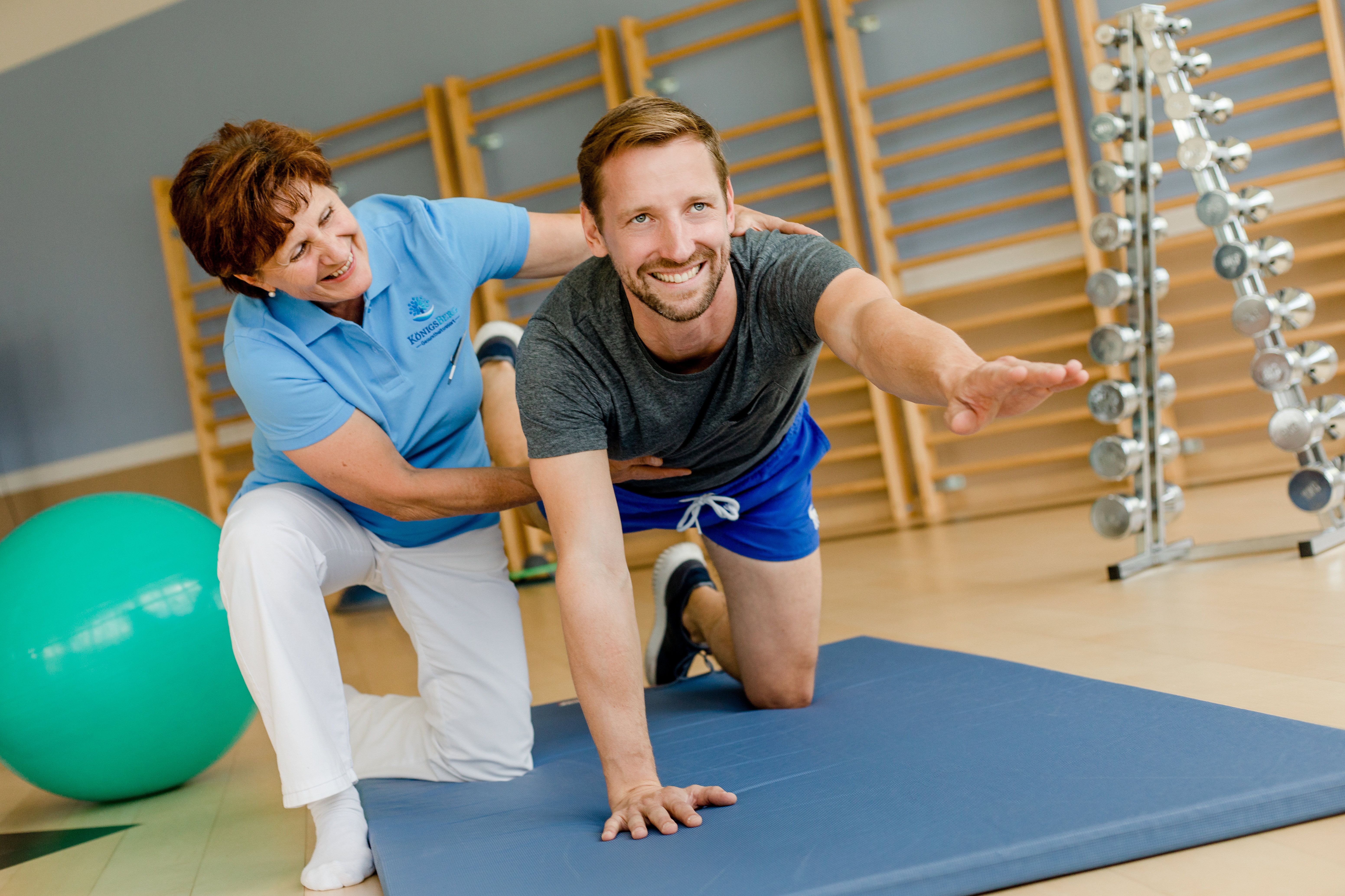 A man does an exercise on a mat, supported by a therapist in a fitness room.
