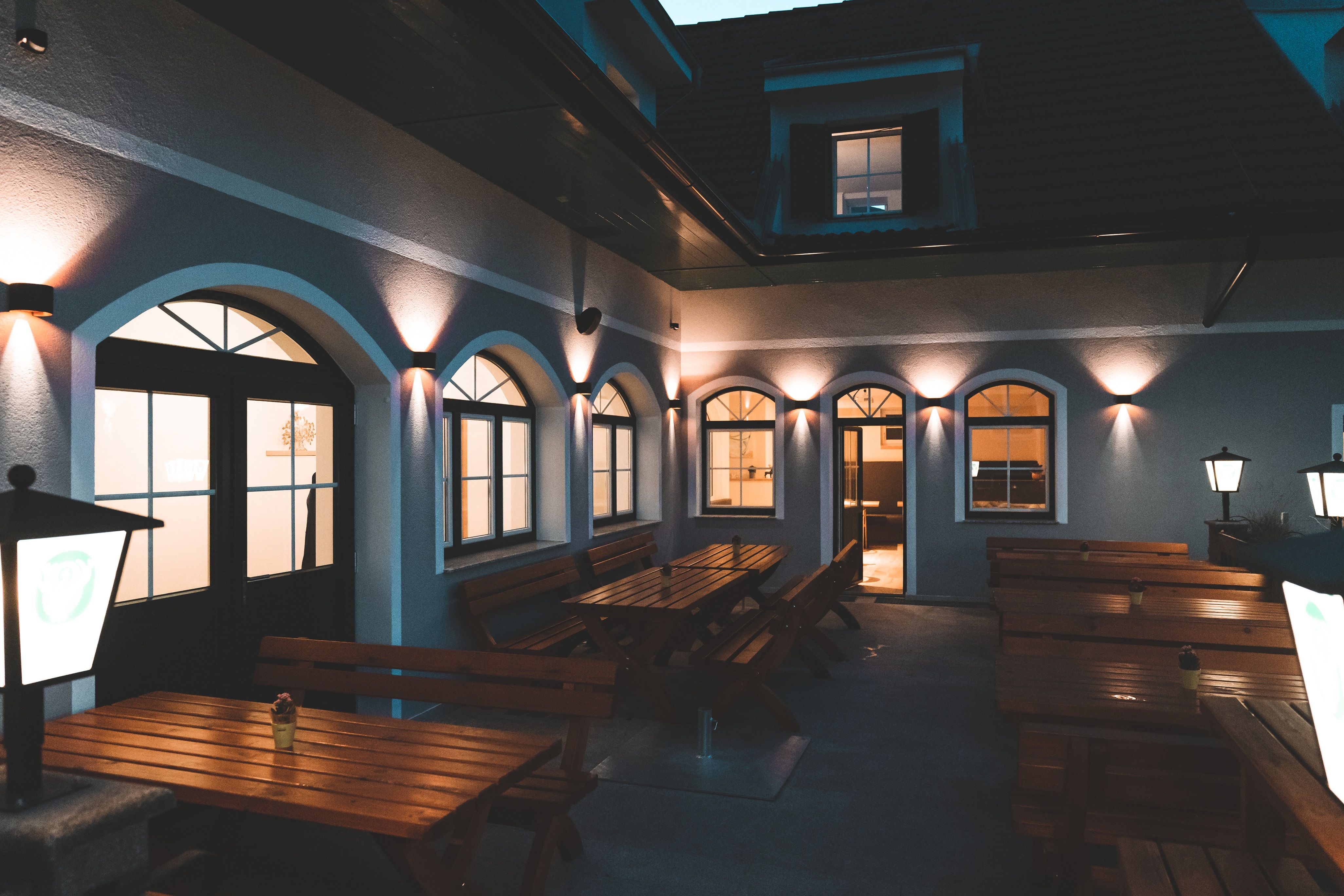 Illuminated courtyard with wooden benches and tables, surrounded by a building with arched windows.