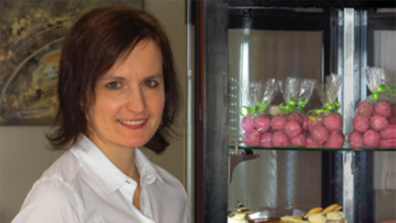 A woman in a white blouse stands smiling next to a display case with packaged pastries.