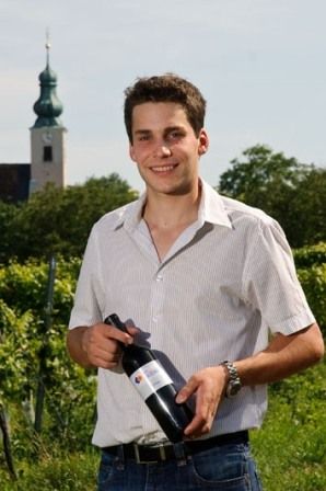 A man stands in a vineyard holding a bottle of wine. A church tower can be seen in the background.