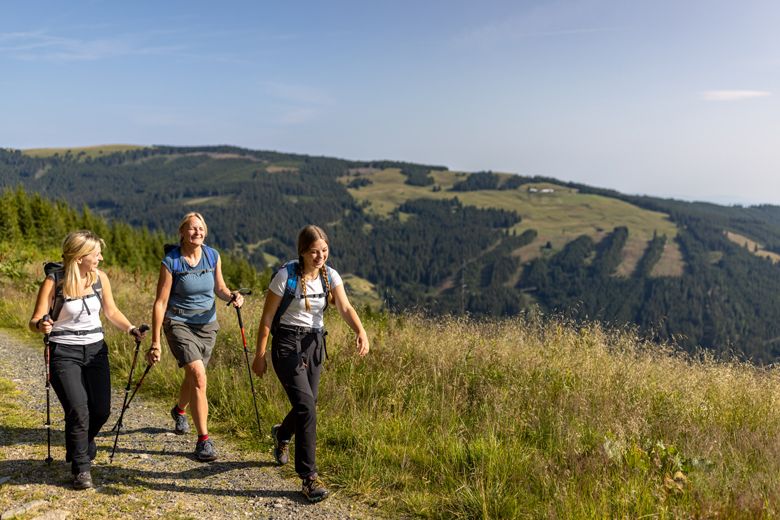 Three women hike along a mountain path with hiking poles and rucksacks, surrounded by green hills and forests.