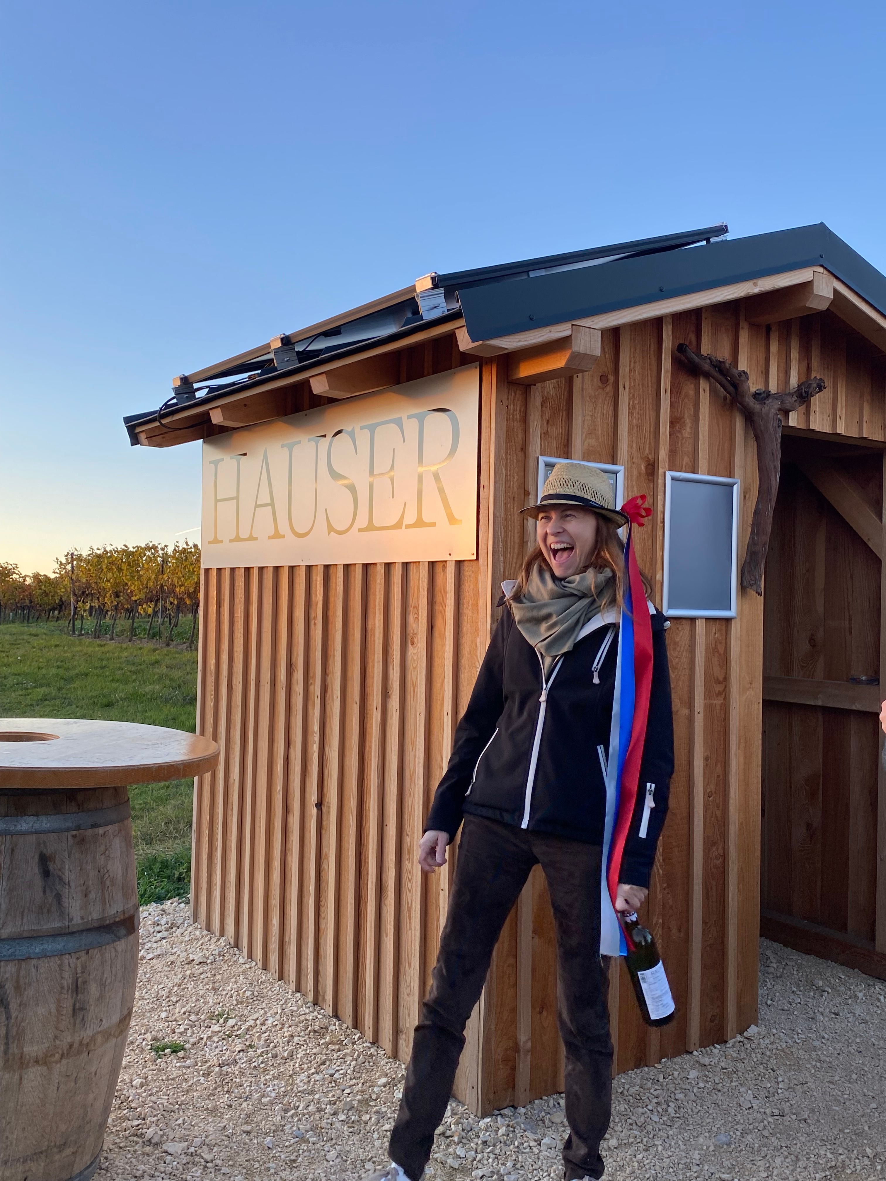 A person with a hat and a bottle stands in front of a wooden hut with the inscription 'Hauser'.