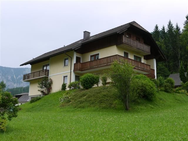 A two-storey house with wooden balconies on a green hill surrounded by trees and meadows.