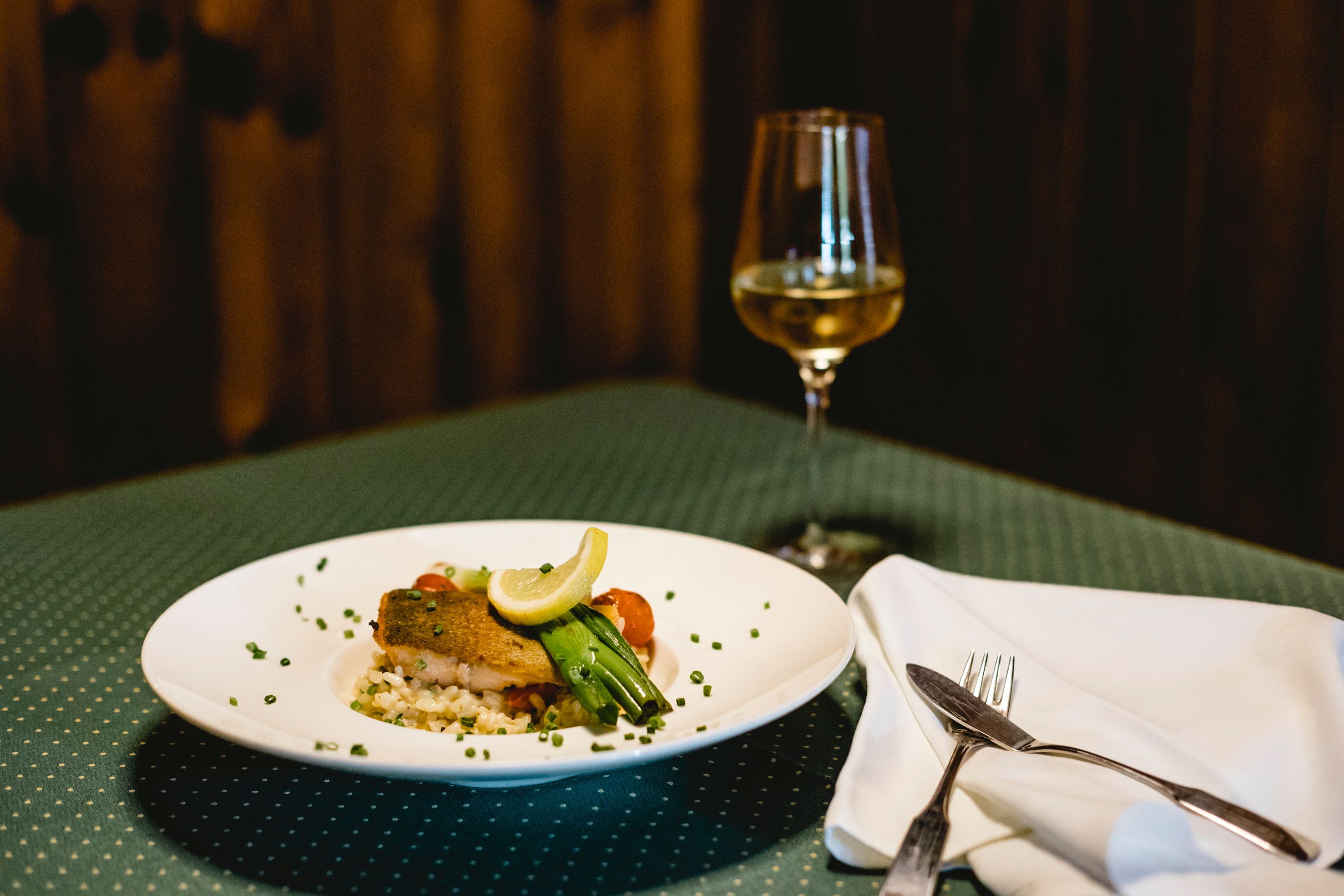 A plate of salmon trout and risotto, garnished with lemon and vegetables, stands on a laid table next to a glass of white wine.