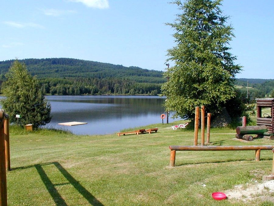 A peaceful lake with a jetty, surrounded by green hills and trees, with a playground in the foreground.