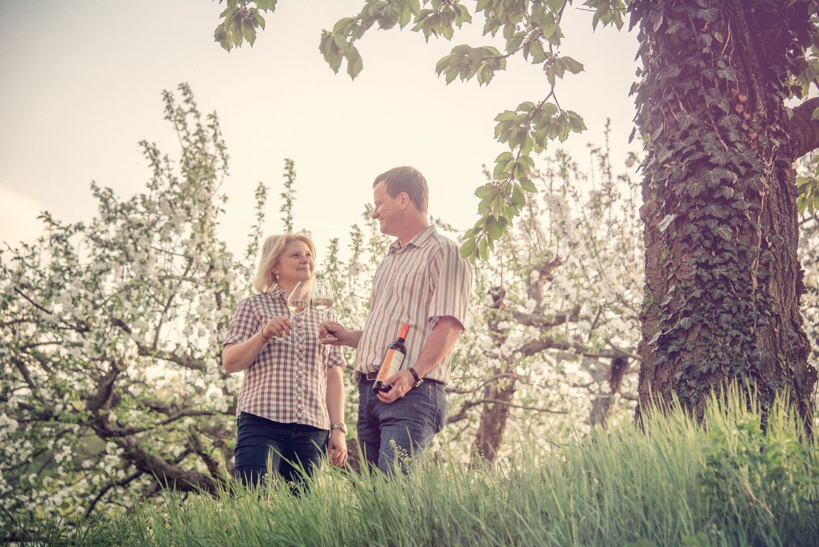 A couple stand in a blossoming orchard and clink glasses of wine while the man holds a bottle of wine.