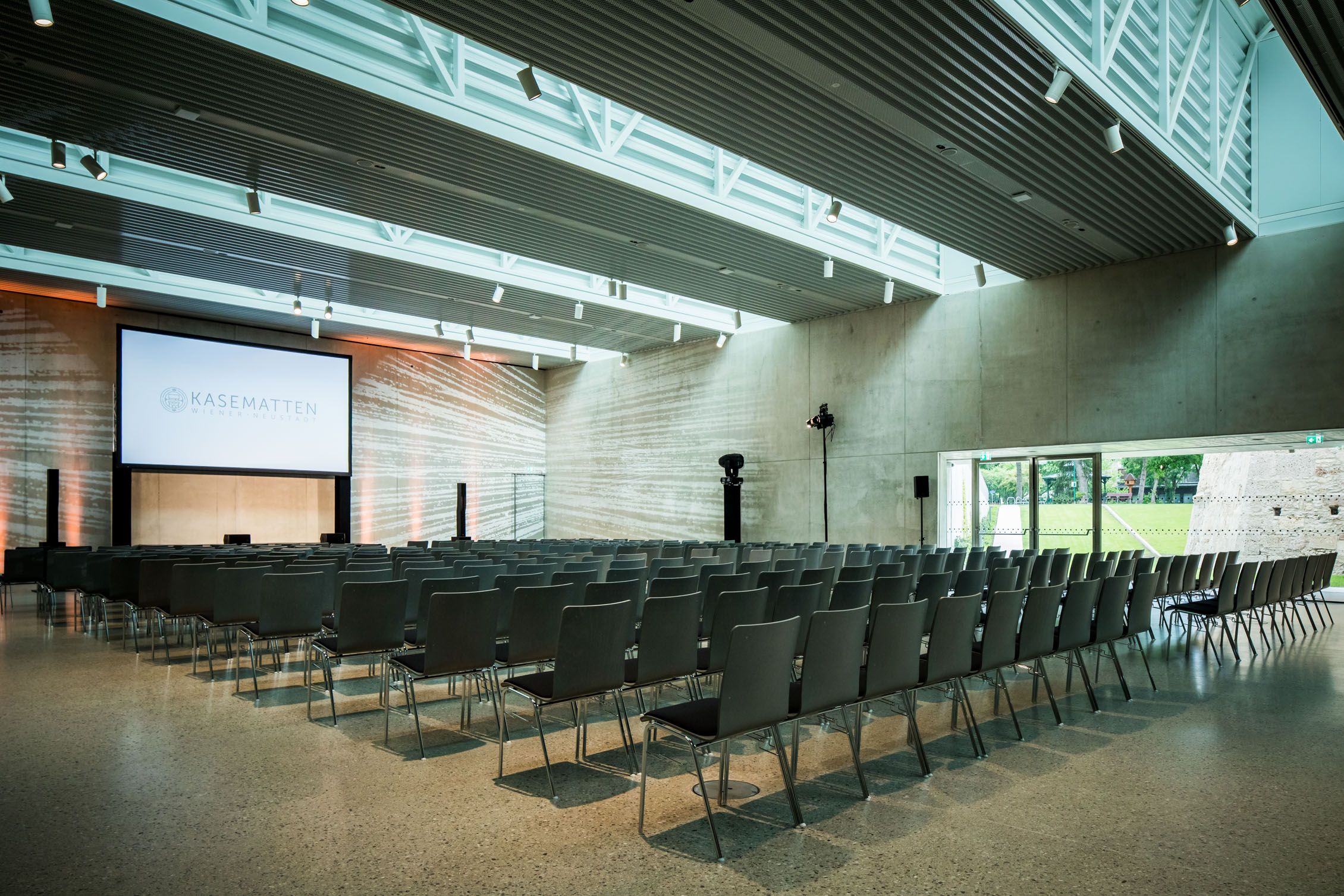 Modern event room with rows of chairs and a large screen, labeled 'Kasematten Wiener Neustadt'.