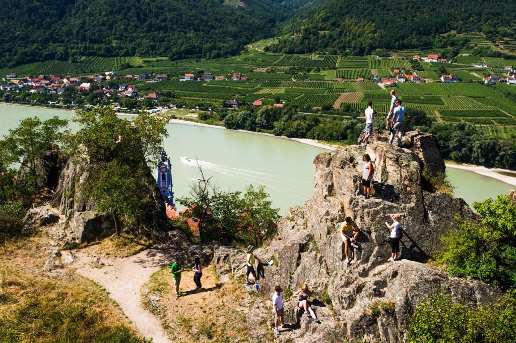 People climbing on rocks with a view of the Danube and vineyards in Dürnstein.
