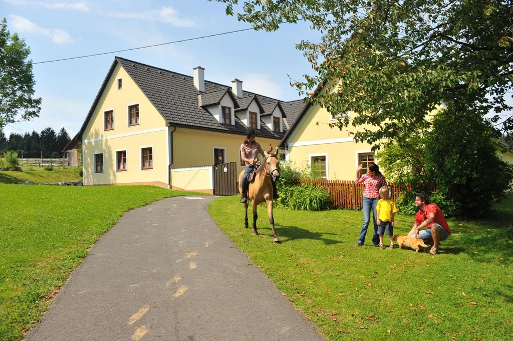 A rider on a horse approaches a family in front of a yellow house on a farm.