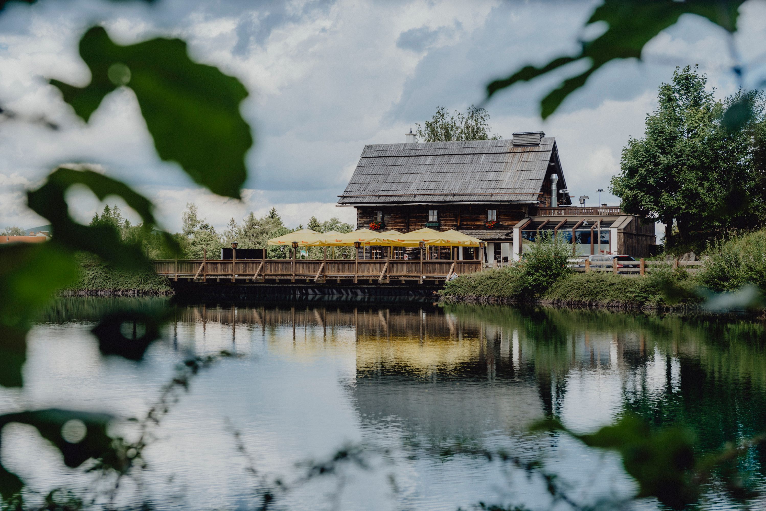 A rustic wooden house with yellow parasols is reflected in a calm lake surrounded by green nature.