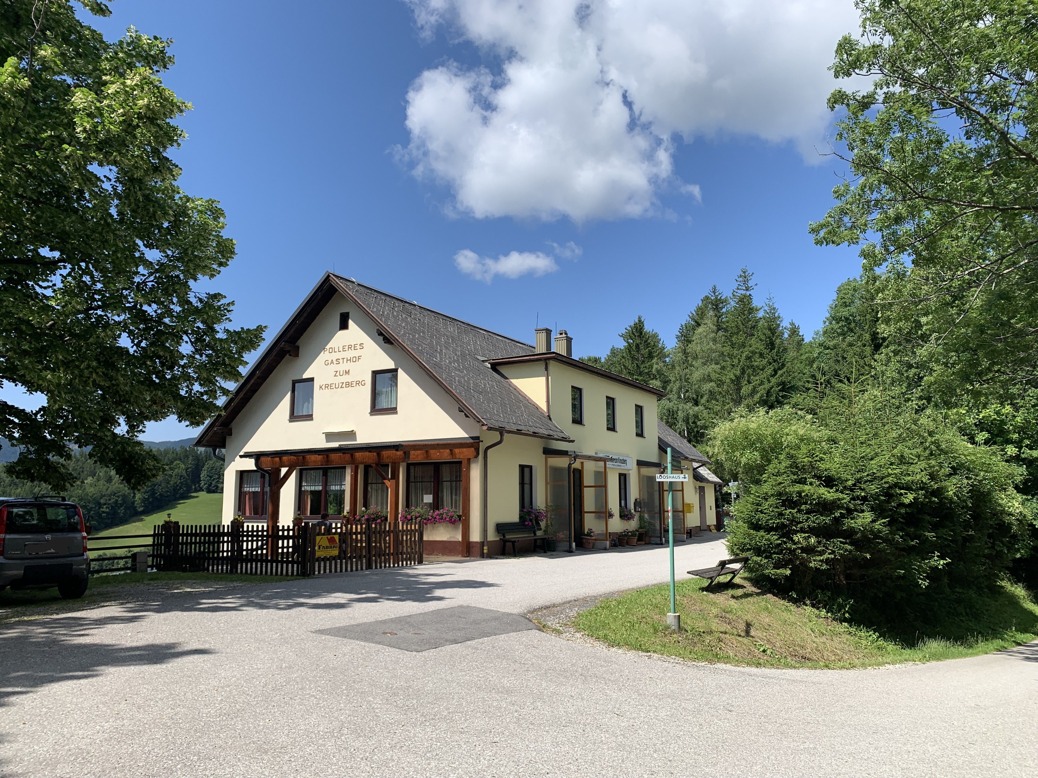 A yellow inn with the inscription 'Polleres Gasthof zum Kreuzberg' surrounded by trees and a blue sky.