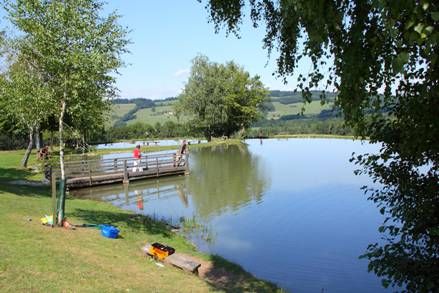 A quiet lake with a jetty and anglers, surrounded by green trees and hills in the background.