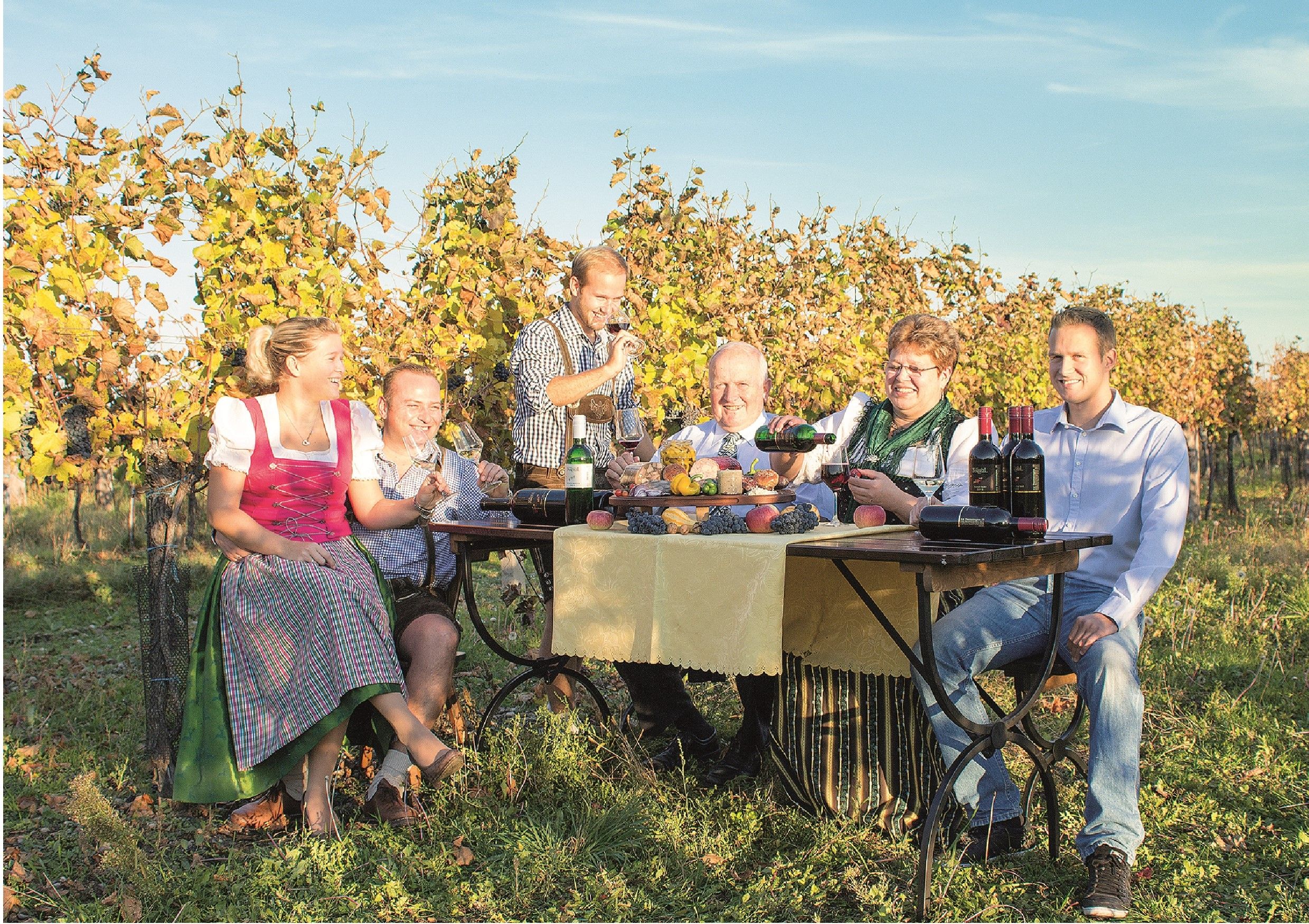 A family sits at a table in the vineyard and enjoys wine and snacks.