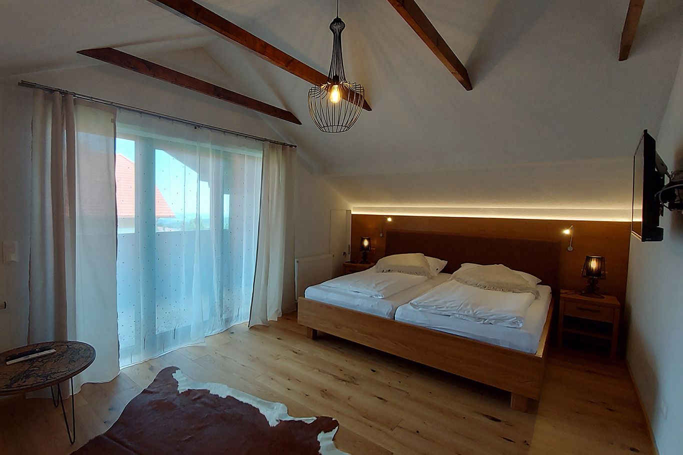 A cozy double room with a wooden floor, a large bed, curtains and a small table. A lamp hangs from the ceiling.