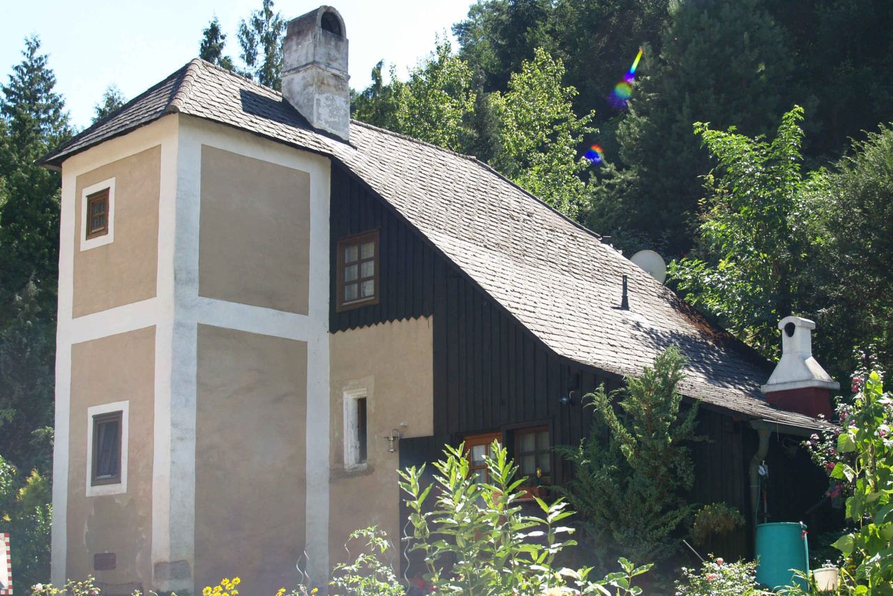 An old house with a small tower and chimney, surrounded by trees and plants.