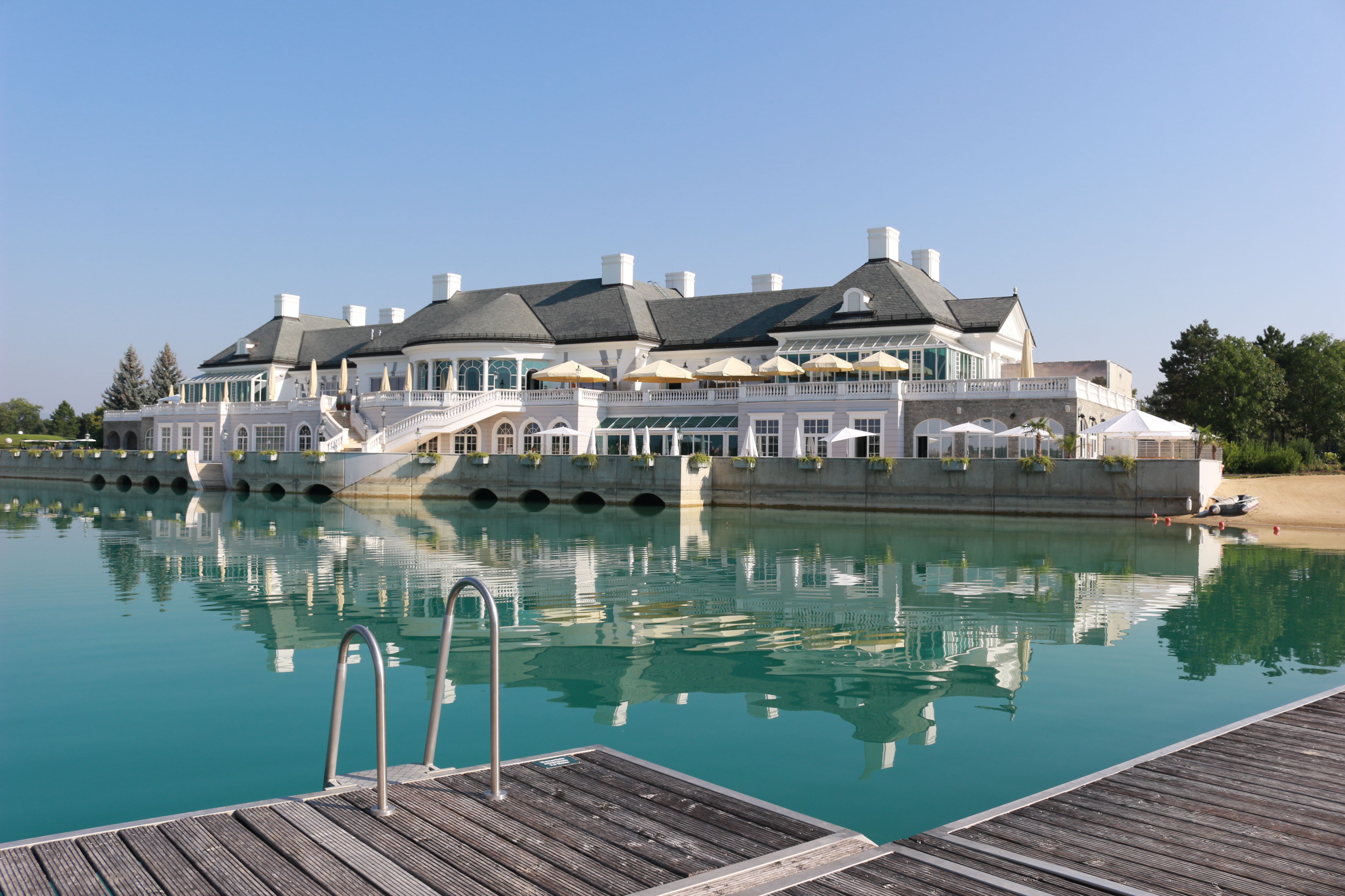 An elegant clubhouse on the lake with a terrace and sunshades, surrounded by clear water and a jetty in the foreground.