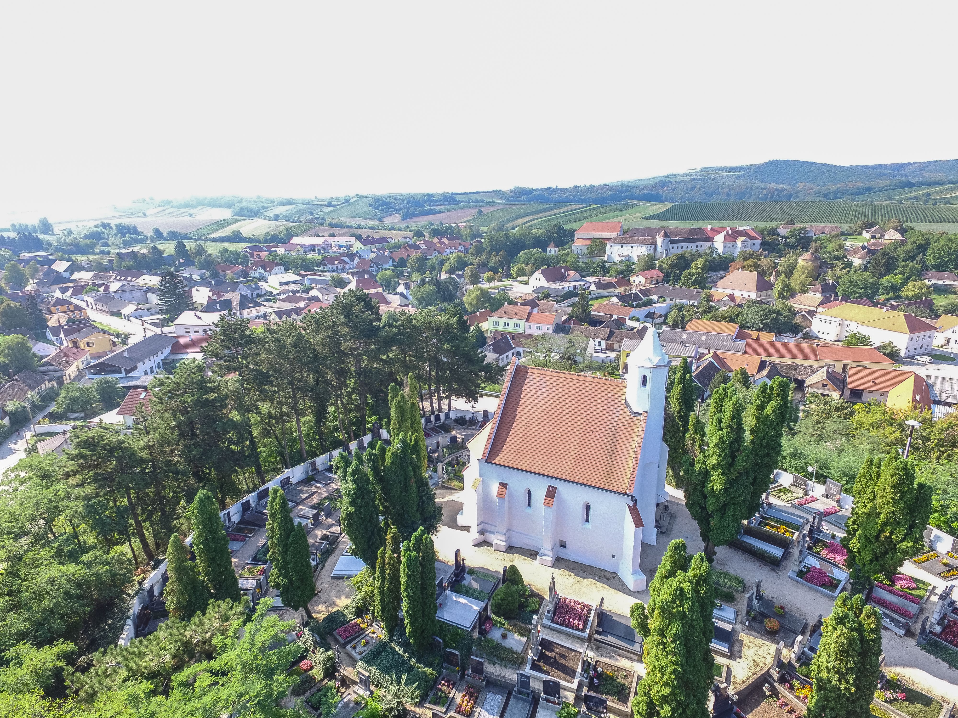 Aerial view of a church with a red roof, surrounded by a cemetery and trees, in a rural setting with houses and hills in the background.