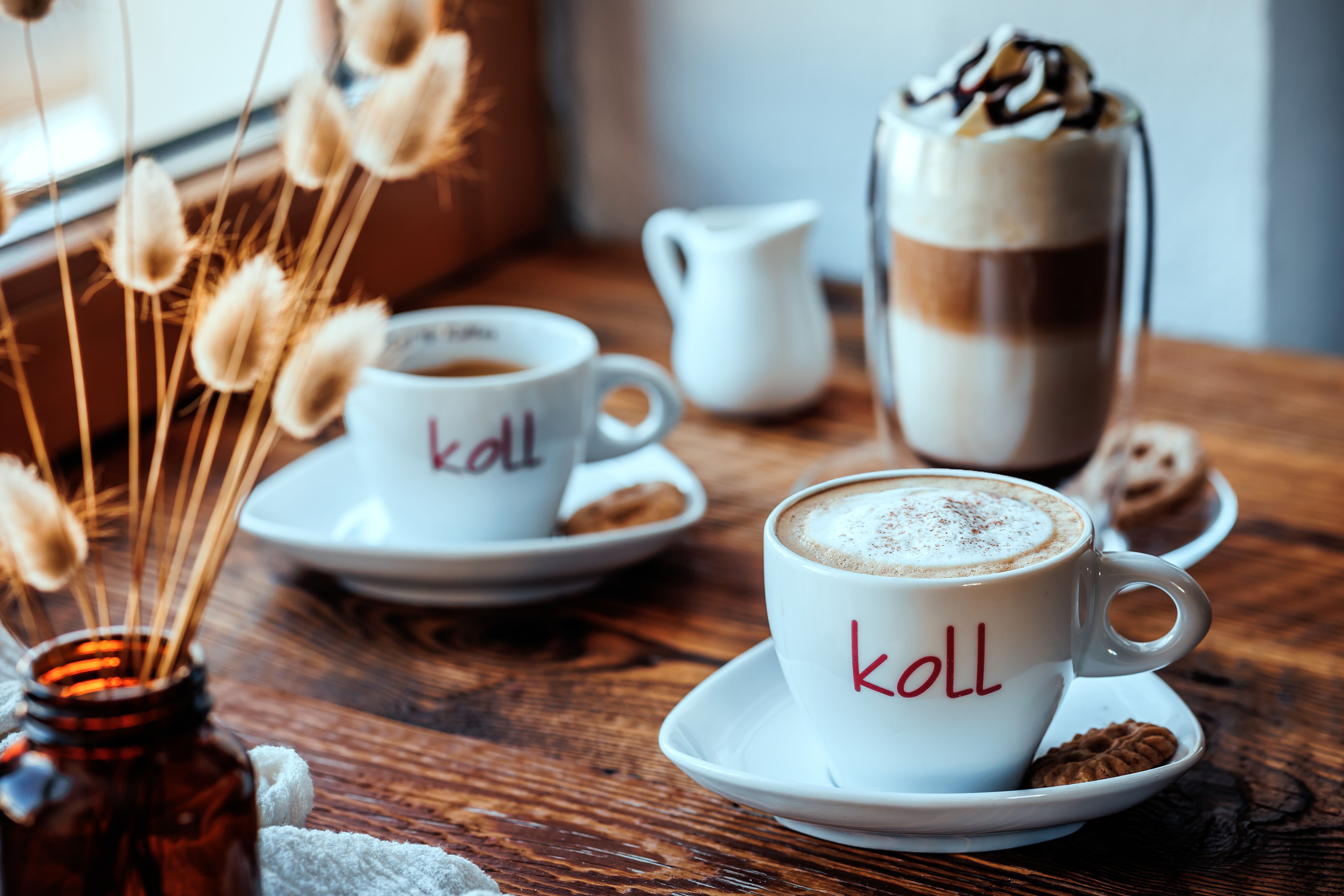 Two cups of coffee on a wooden table, one with milk foam, a glass of latte macchiato and a jug next to it.