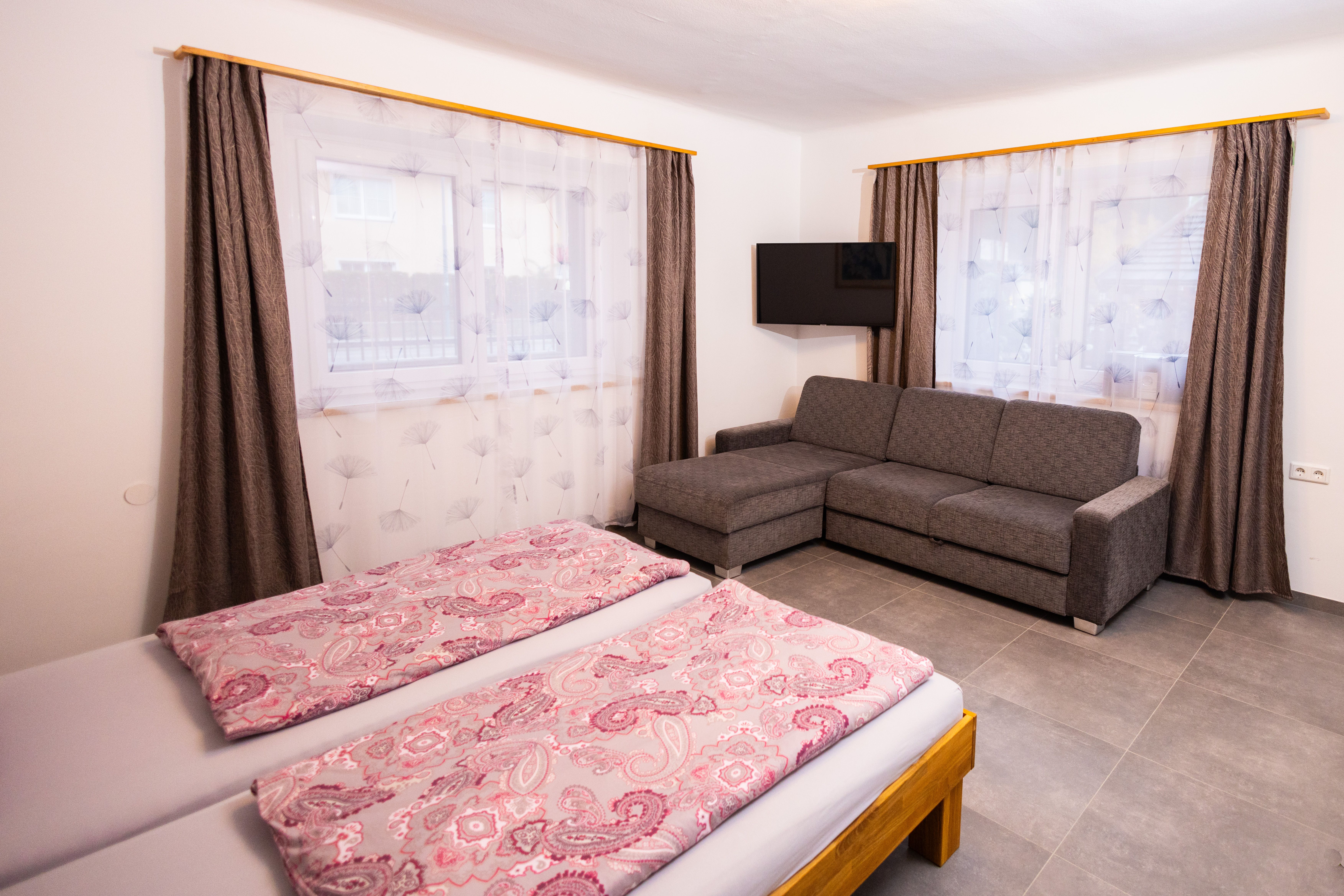 One bedroom with double bed, gray sofa, TV and two windows with curtains.