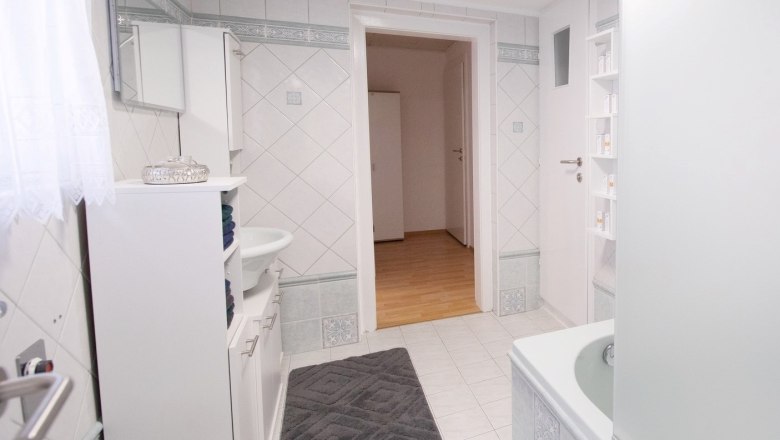 Modern bathroom with white tiles, bathtub, washbasin and cabinet. Door leads to a hallway with wooden floor.